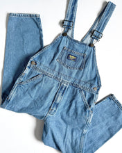 Load image into Gallery viewer, A pair of light blue denim work overalls laid flat on a white background, with the legs folded over the bib. The overalls feature adjustable shoulder straps with metal buckles, a front patch pocket with a small yellow and blue brand label, and front flap pockets.
